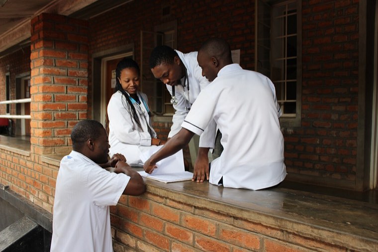 Three healthcare workers in white coats, two with stethoscopes, review a patient file outside a hospital building